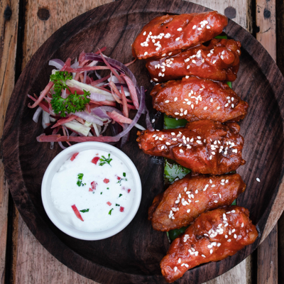 A top view of some neatly lined up saucy hot wings topped with sesame seeds, served with a side of blue cheese dip and garnish.